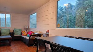 Una habitación con una mesa de comedor, sillas, un sofá con cojines, una ventana que da a árboles y un suelo de baldosas.
