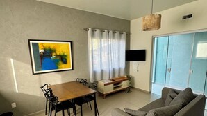 Ett vardagsrum med en grå soffa, ett matbord i trä med stolar, en tv som är monterad på väggen och ett fönster med gardiner.