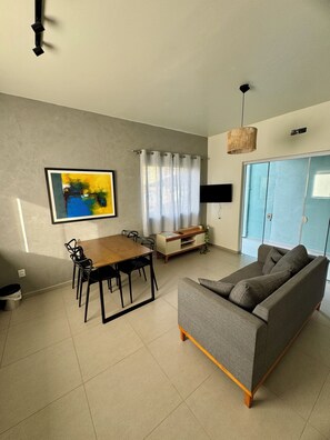 Ein Wohnzimmer mit grauem Sofa, einem Holzespeisetisch mit Stühlen, einem an der Wand befestigten Fernseher und einem Fenster mit Vorhängen.