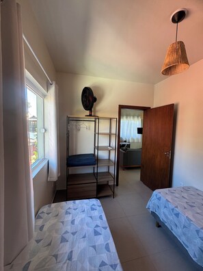 Ein kleines Zimmer mit zwei Betten, einem Standventilator, einem Holzschrank und einem Fenster mit Vorhängen.