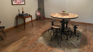 Un comedor con una mesa redonda, cuatro taburetes, una mesita auxiliar con un juego de damas, un cuadro en la pared y una puerta con un cerrojo.