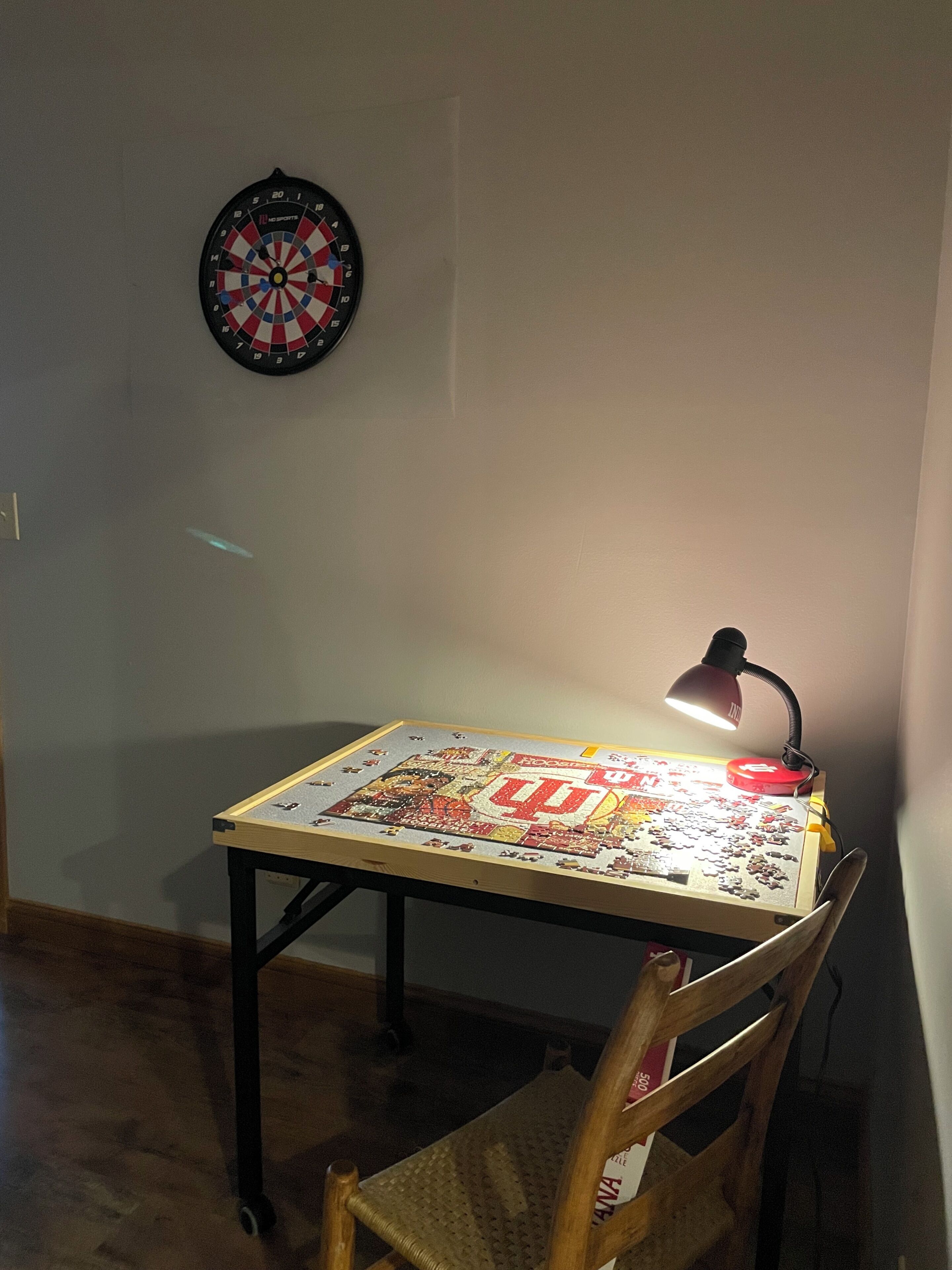 A table with a puzzle, a desk lamp, a dartboard, and a chair.