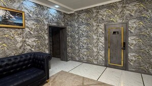 A lobby with a chandelier, a black sofa, marble walls, and a door with a number plate. - Royal Hotel Navoi (Navoiy)