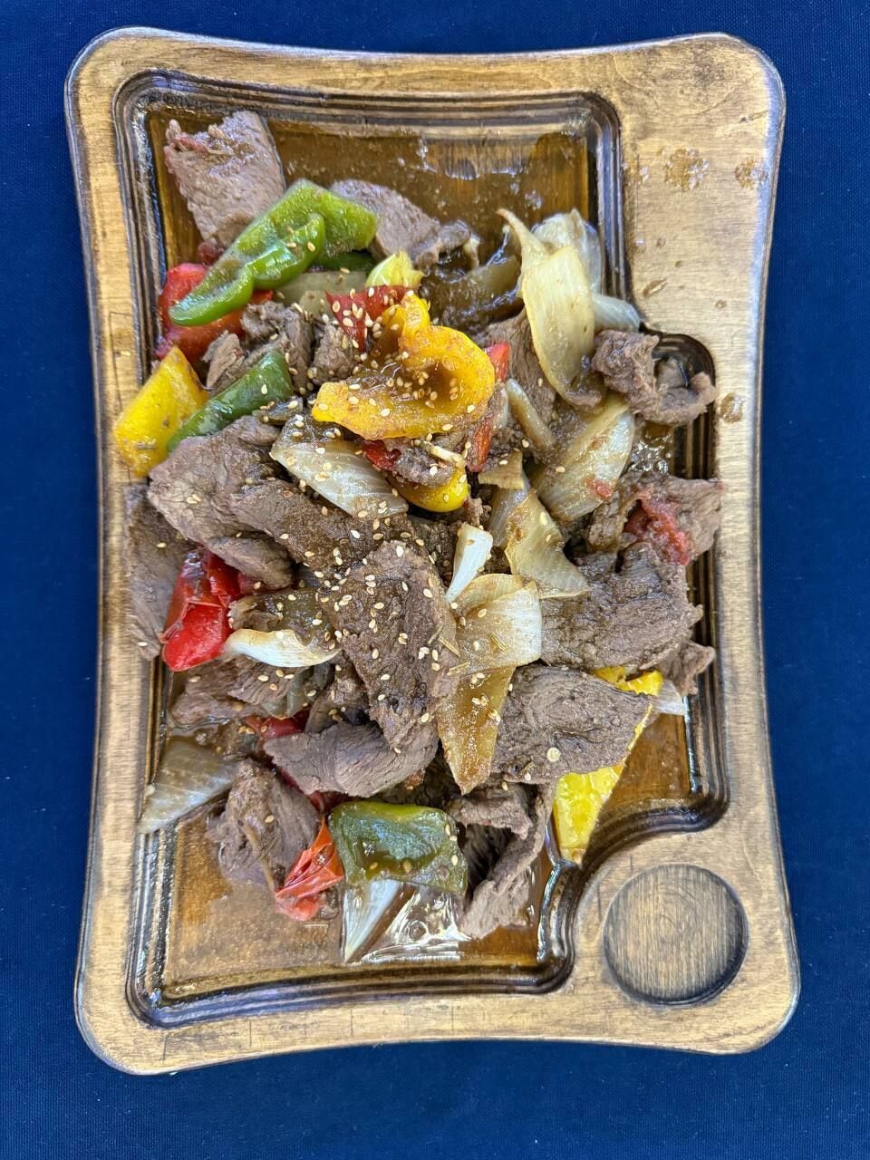 A square-shaped serving dish with a wooden handle, containing a meal of sliced beef, bell peppers, and onions, garnished with sesame seeds.