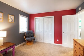 A room with a grey wall, red accent wall, white doors, a wicker chair, a wooden dresser, and a table with a lamp. - Tideland Landing - Your 3 bedroom Ocracoke escape awaits (Ocracoke)