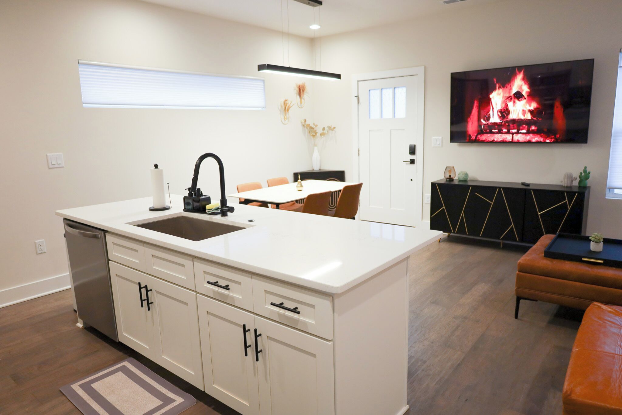 A modern kitchen with a central island, a dining area with chairs, a flat-screen TV mounted above the island, and a black console table with decorative items.