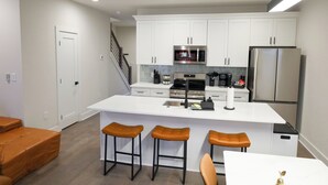 Cocina moderna con armarios blancos, un refrigerador de acero inoxidable, microondas y cafetera. Hay tres taburetes en la barra central.