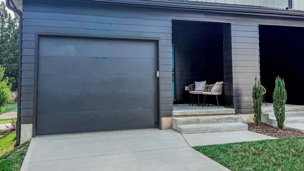 Una casa moderna de dos pisos con puerta de garaje gris oscuro, un porche cubierto con mobiliario exterior y un césped bien cuidado.