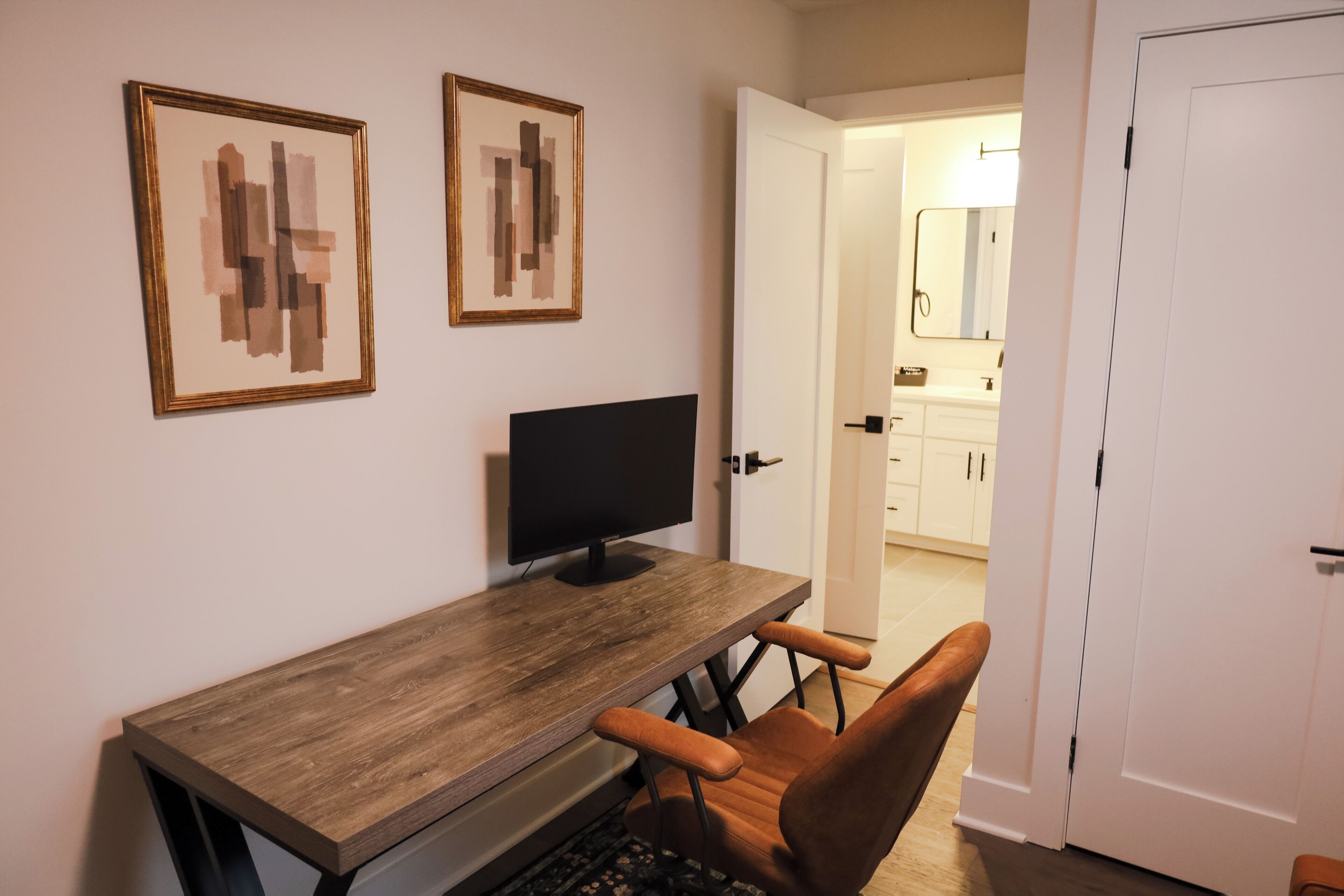 A room with a wooden desk, a black monitor, two framed abstract paintings, and a brown chair.