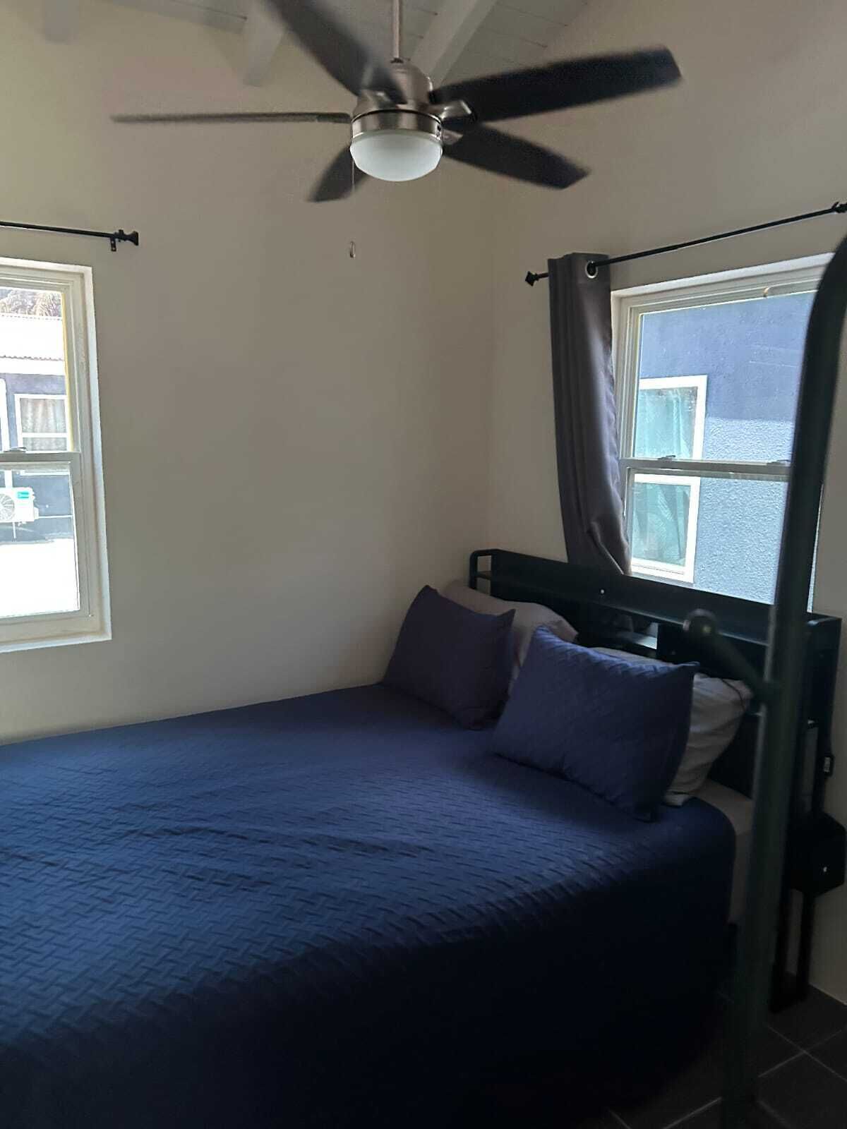 Una cama individual con una colcha y almohadas azul oscuro, un ventilador de techo y una ventana con cortinas.