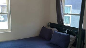 Una cama individual con una colcha y almohadas azul oscuro, un ventilador de techo y una ventana con cortinas.