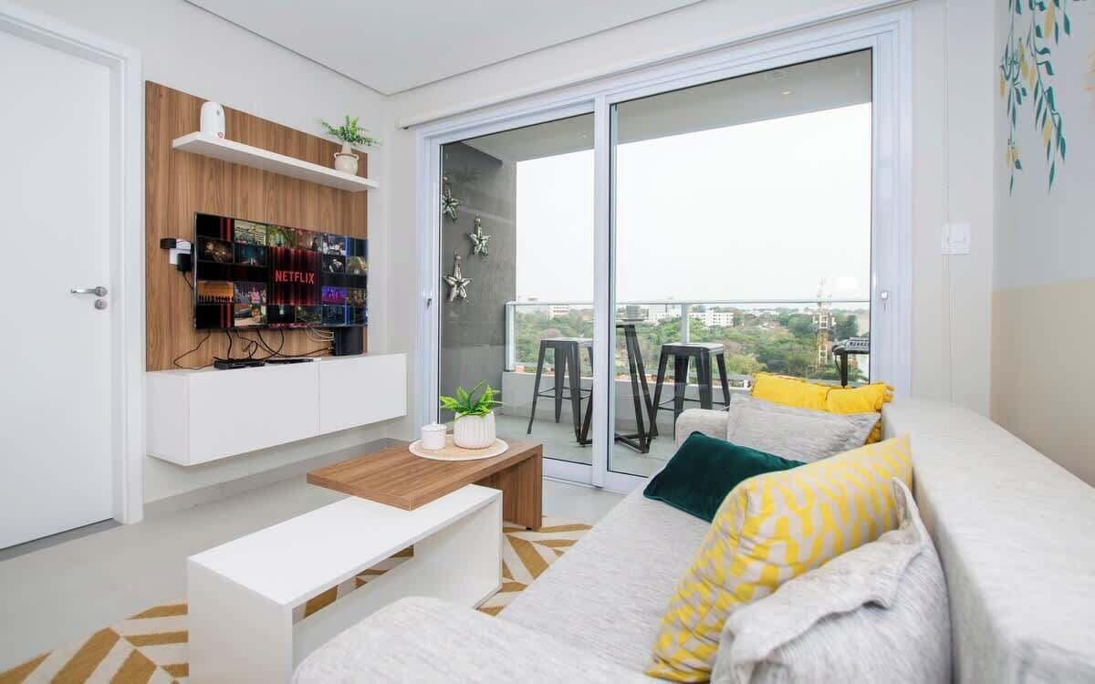 A modern living room with a sofa, coffee table, TV mounted on a wooden shelf, and a sliding glass door leading to a balcony.