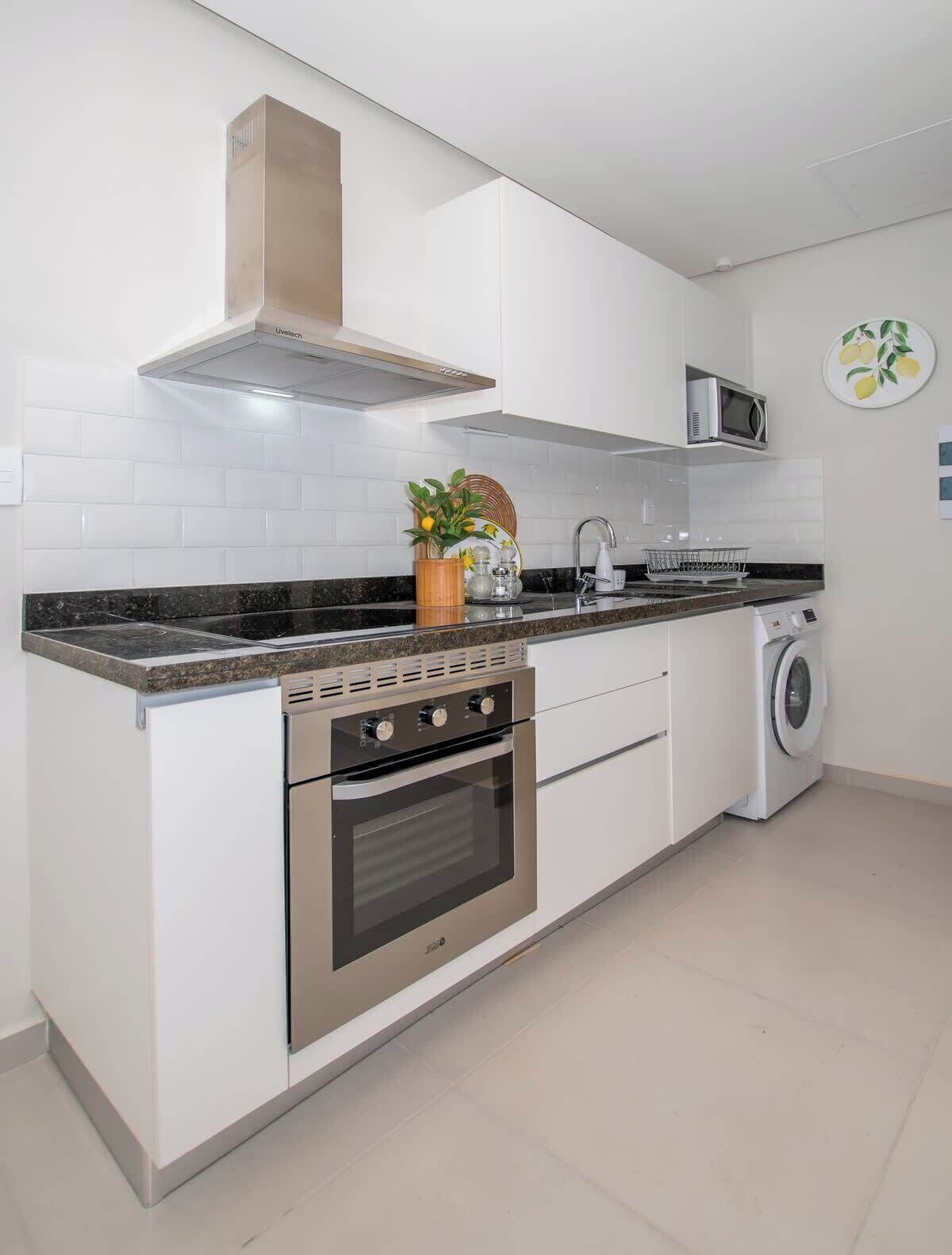 A modern kitchen with a built-in oven, microwave, and washing machine, white cabinetry, and a tiled backsplash.