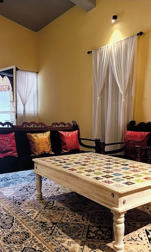 A room with a wooden table, a patterned rug, a sofa with cushions, and a window with curtains. - Heritage Haven (Ashwem)