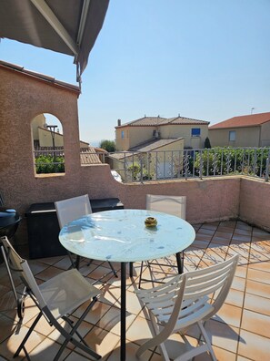 A patio with a round table and chairs, a grill, and a view of neighboring buildings. - Beachfront apartment, very large terrace, pool, air conditioning, parking (Le Barcarès)