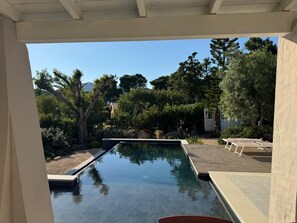 A swimming pool with a wooden deck and lounge chairs, surrounded by trees and a clear sky. - House Among the Olive Trees (villasimius)