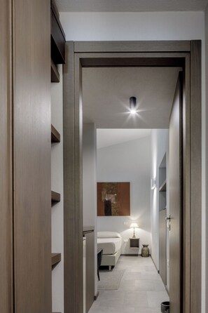 A narrow hallway leading to a bedroom with a bed, bedside table, lamp, and a wall-mounted artwork. - House Among the Olive Trees (villasimius)