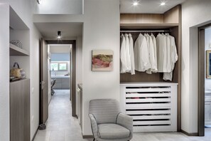 A modern interior with a hallway leading to a kitchen, a seating area with a patterned chair, and a clothing storage unit with hanging garments. - House Among the Olive Trees (villasimius)