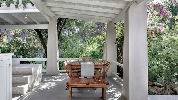 Une terrasse extérieure couverte, avec une table et des chaises en bois, entourée de verdure et d’un escalier en pierre.