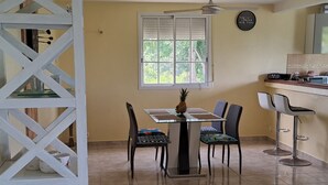 A dining area with a table, chairs, a pineapple, a window, and a kitchen area with a refrigerator and a clock. - Joss location  971 (Le Gosier)