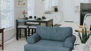 A modern living room with a blue sofa, a dining area with a table and chairs, a kitchen with white cabinets, and a large window. - Modern 2BR • Gated Parking • 10 Min to Downtown (Detroit)