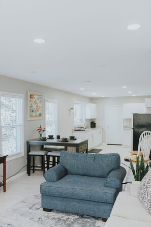 A modern living room with a blue sofa, a dining area with a table and chairs, a kitchen with white cabinets, and a large window. - Modern 2BR • Gated Parking • 10 Min to Downtown (Detroit)