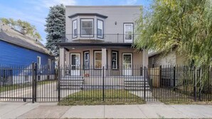 A two-story house with a front porch, a bay window, and a fenced yard. - Modern 2BR • Gated Parking • 10 Min to Downtown (Detroit)
