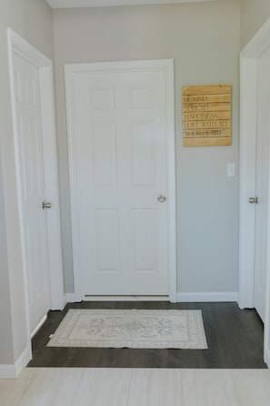 Un pasillo con una puerta blanca, un letrero de madera con texto inspirador y una alfombra estampada sobre un piso oscuro.