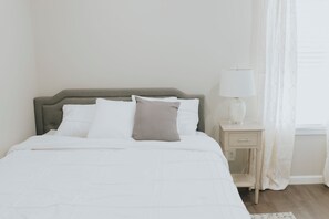 A bed with white bedding and a grey headboard, a bedside table with a lamp, and a window with sheer curtains. - Modern 2BR • Gated Parking • 10 Min to Downtown (Detroit)