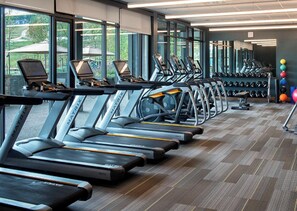 Un gimnasio con cintas de correr, un estante lleno de mancuernas y grandes ventanales con vistas al exterior.