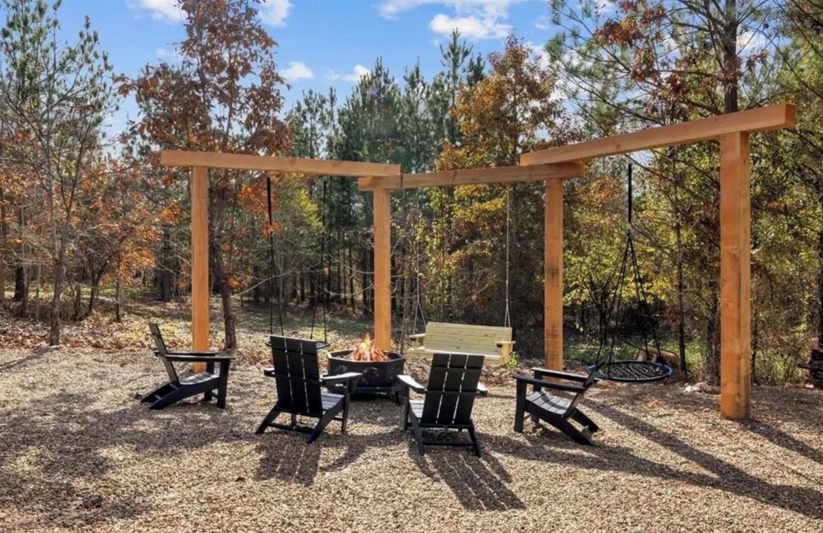 Une pergola en bois avec des sièges, un foyer et une balançoire, le tout situé dans une zone boisée.