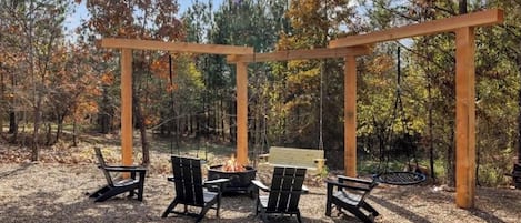 Una pérgola de madera con asientos, un brasero y un columpio en un área boscosa.