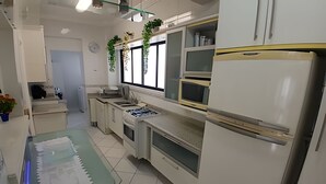 Een compacte keuken met witte kasten, een magnetron, een fornuis en een koelkast. Er is een raam met planten op de vensterbank en een kleine tafel met een keukenrolhouder.