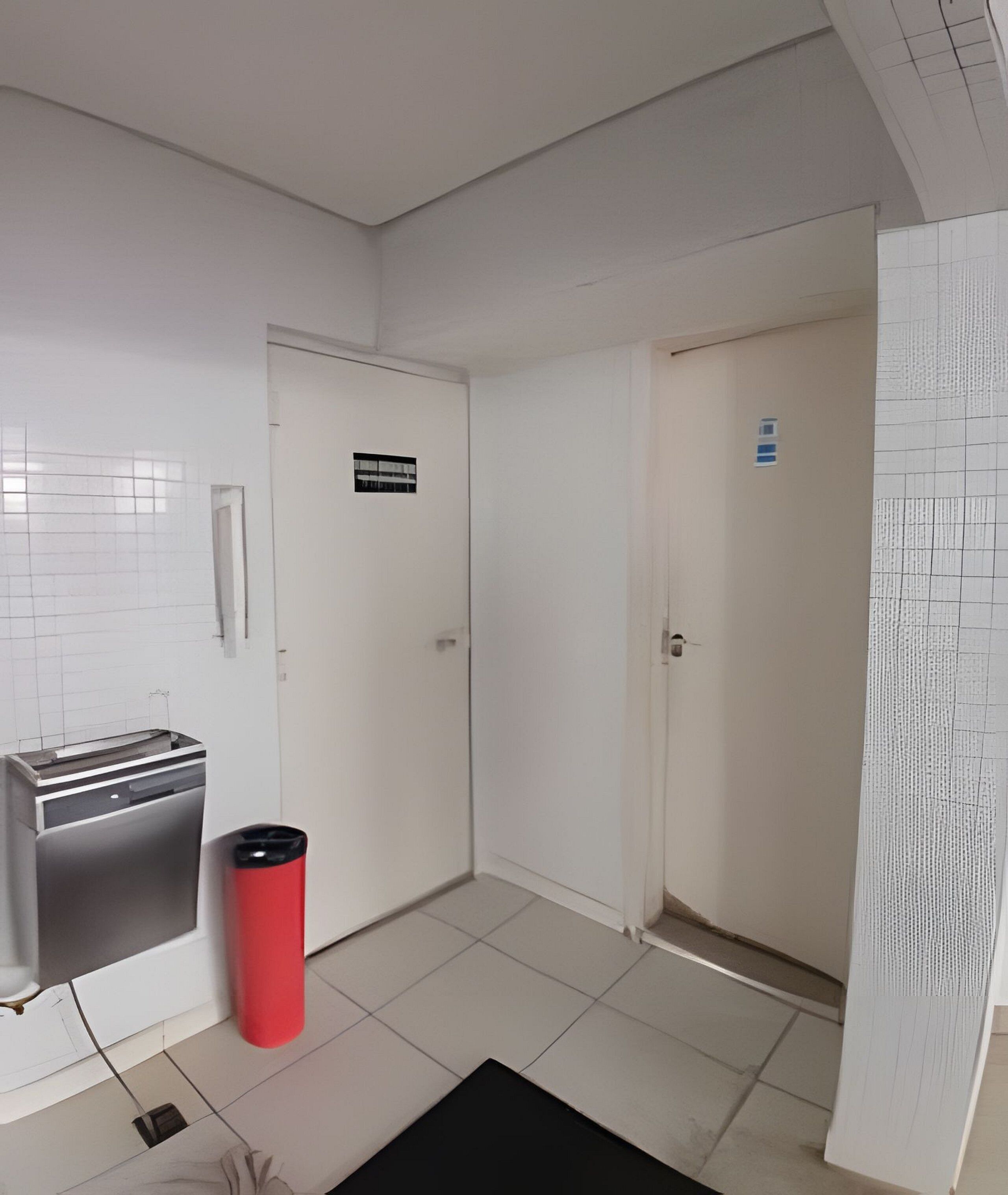 A hallway with white walls, a tiled floor, and a red trash bin. There are two doors, one with a sign above it.