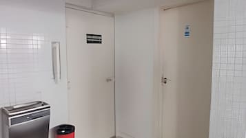Un pasillo con paredes blancas, piso de baldosas y un bote de basura rojo. Hay dos puertas, una con un letrero encima.