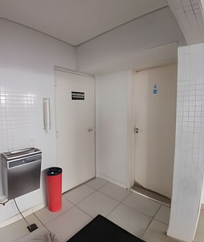 Ein Flur mit weißen Wänden, einem gefliesten Boden und einer roten Mülltonne. Es gibt zwei Türen, über einer davon ein Schild.
