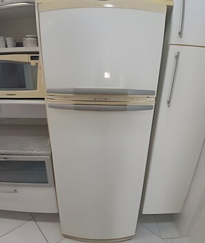 Un refrigerador blanco con un compartimento congelador en la parte superior, ubicado en una cocina con gabinetes blancos y un microondas.