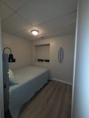 Ein kleines Schlafzimmer mit einem Bett, einem Fenster mit Jalousien, einer Wandleuchte und einer Surfbrettherdekorations.