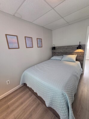A bedroom with a bed, wooden headboard, wall-mounted light, and three framed pictures. - At the mouth of Ocean Blvd short walk to the Beach Unit has been newly decorated (Hampton)