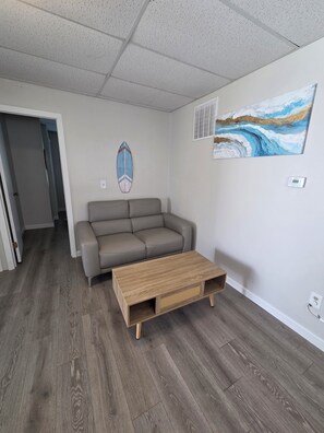 Ein Wohnzimmer mit grauem Sofa, einem Holztisch, einer Surfbrett-Dekoration und einem Wellenbild an der Wand.