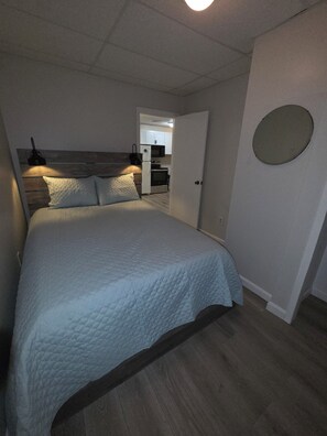 Ett sovrum med en säng, ett sängbord, en väggmonterad lampa och en dörr som leder till ett litet kök.