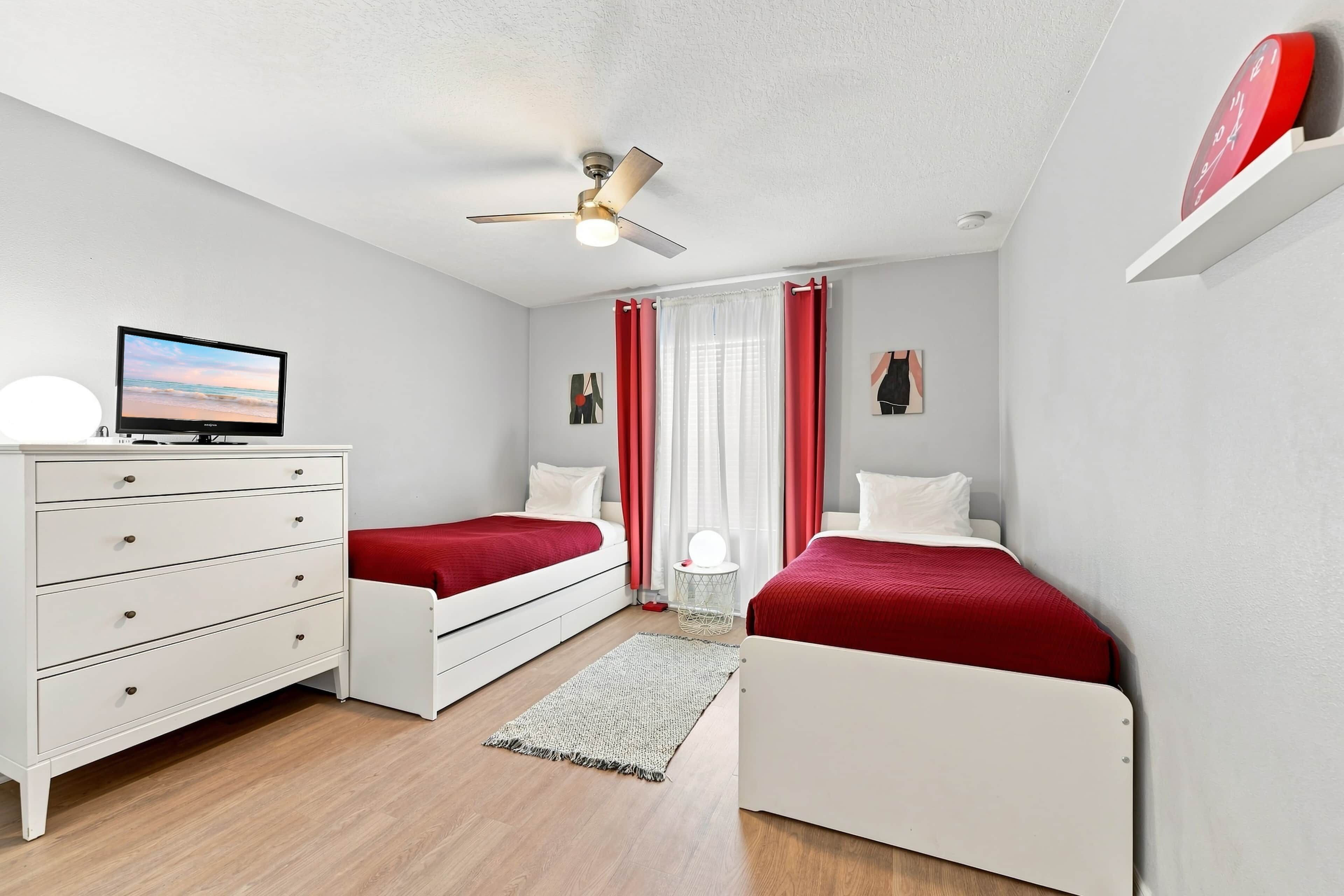 Quarto com duas camas de solteiro, cômoda branca, ventilador de teto, televisão e janela com cortinas.