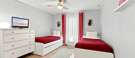 A room with two single beds, a white dresser, a ceiling fan, a TV, and a window with curtains.