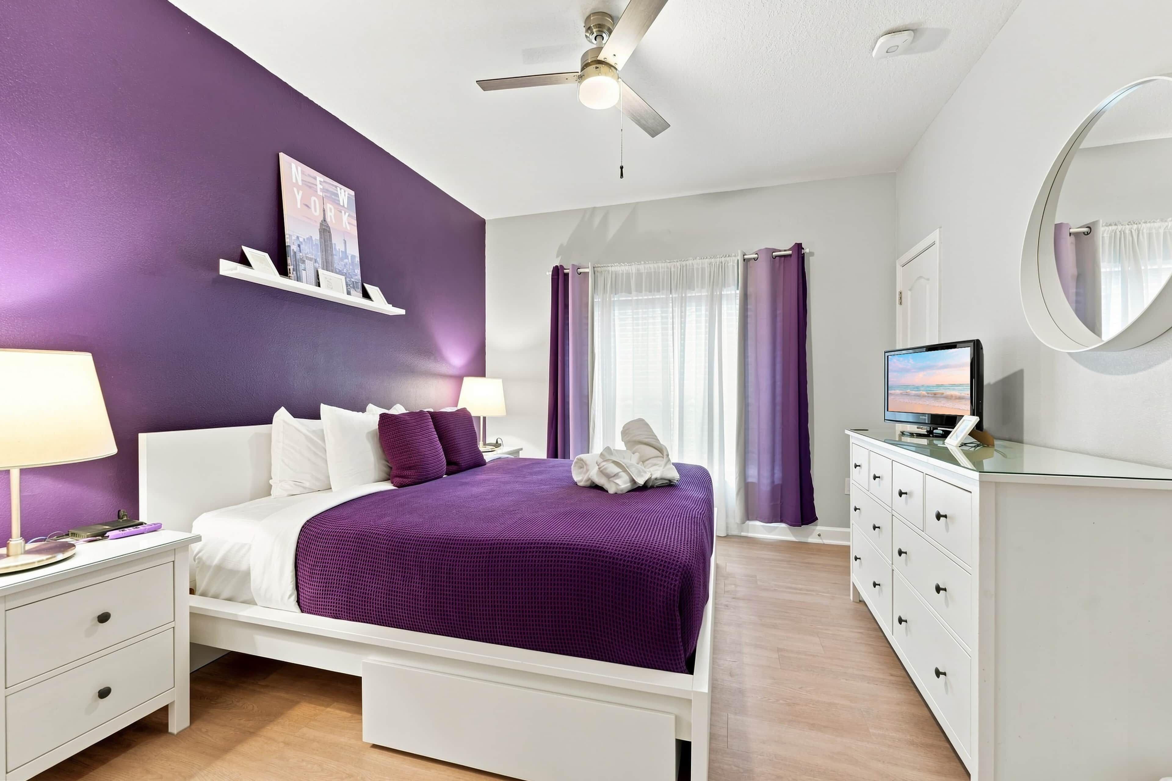 Een slaapkamer met een bed, nachtkastje, dressoir, plafondventilator, raam met gordijnen en een aan de muur bevestigde televisie.