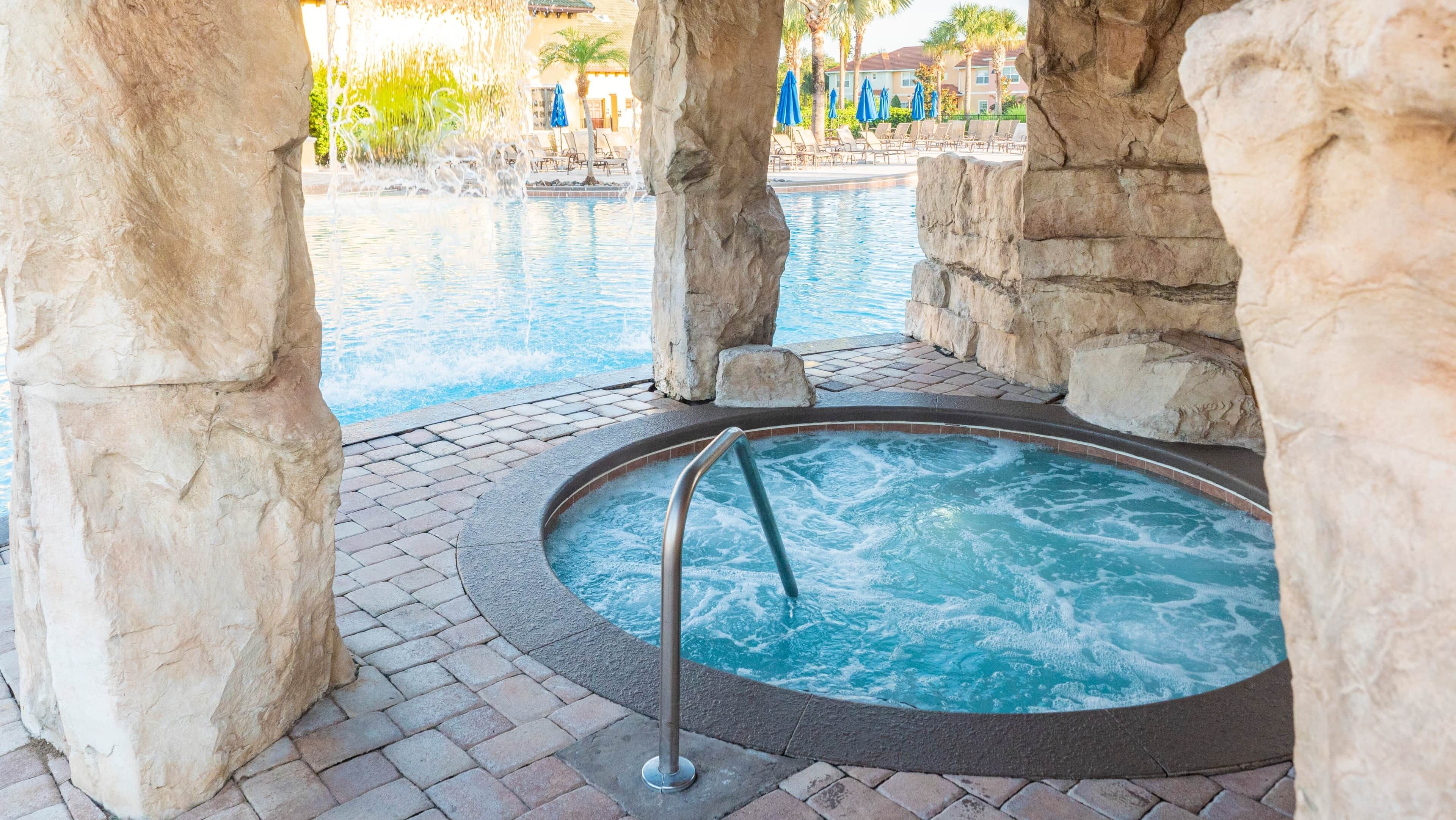 A hot tub area with stone pillars, a pool, and lounge chairs in the background.