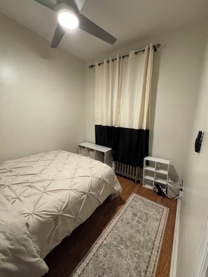 Ein Schlafzimmer mit einem Bett, einem Deckenventilator, einem Heizkörper, einer kleinen weißen Ablage und einem Teppich.