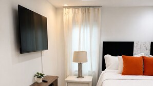 A modern bedroom with a flat-screen TV, a bedside table with a lamp, a bed with white bedding and orange pillows, and a wooden console unit. - 3BR House Near Mercedes Stadium Sleeps 8 w/ free Parking, A/C, Laundry, & Gazebo (Atlanta)