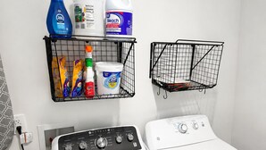 A laundry area with a front-loading washer and dryer, a shelf with cleaning supplies, and a black wire basket. - 3BR House Near Mercedes Stadium Sleeps 8 w/ free Parking, A/C, Laundry, & Gazebo (Atlanta)