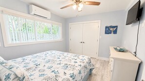 Un dormitorio con cama, ventilador de techo, televisor y ventana con persianas.