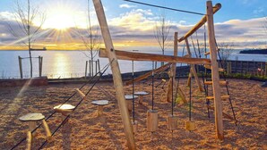 A wooden swing set and climbing structures on a sandy area with a sunset view over water.